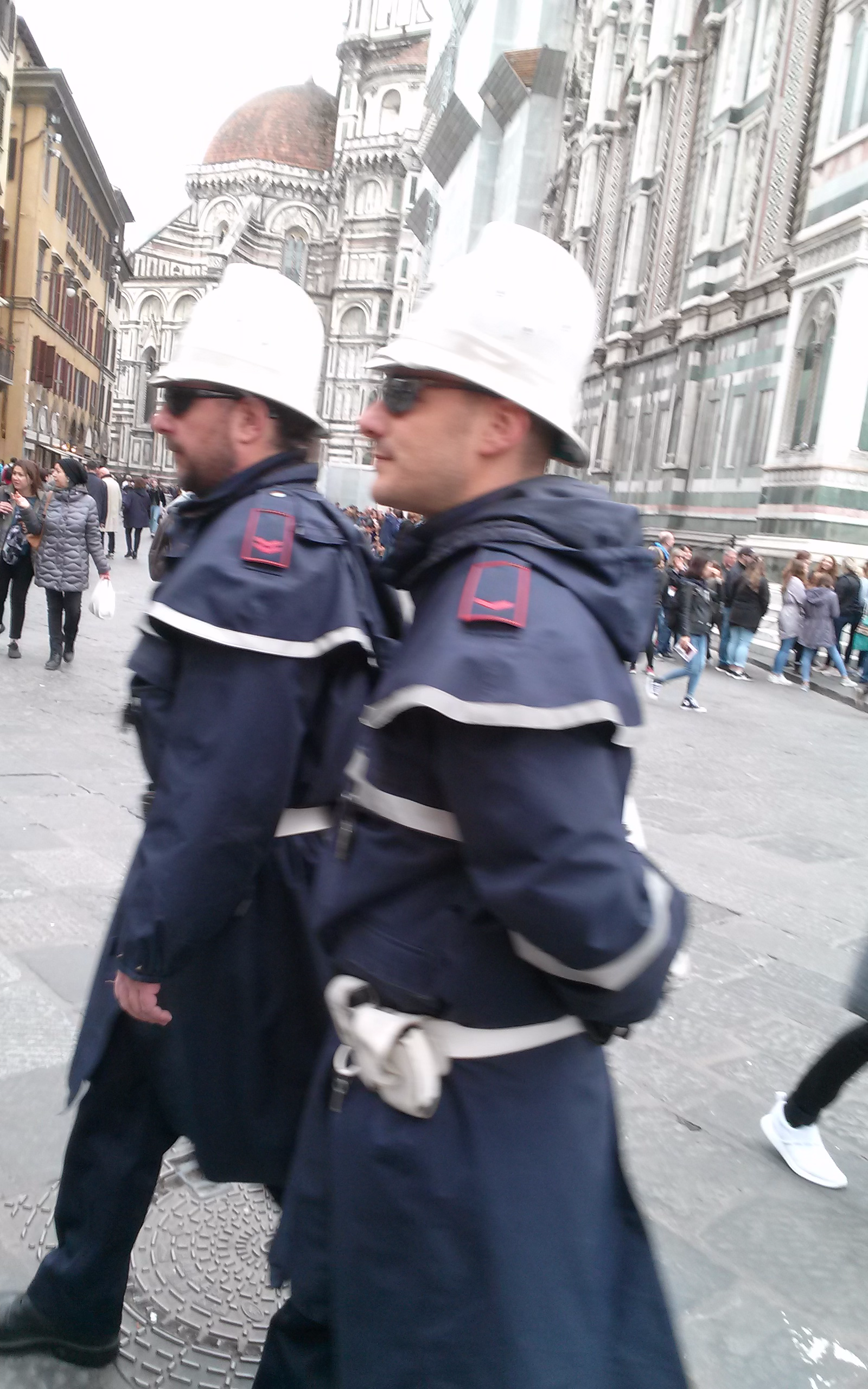 Two men in blue jackets and white hats walking down a street.